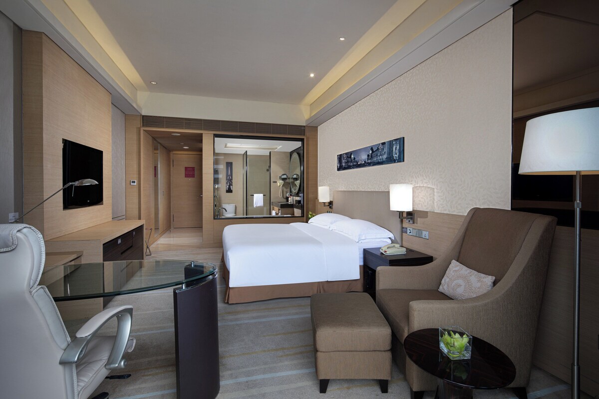 A hotel room with a bed, a desk, a chair, a small table, a lamp, and a TV.