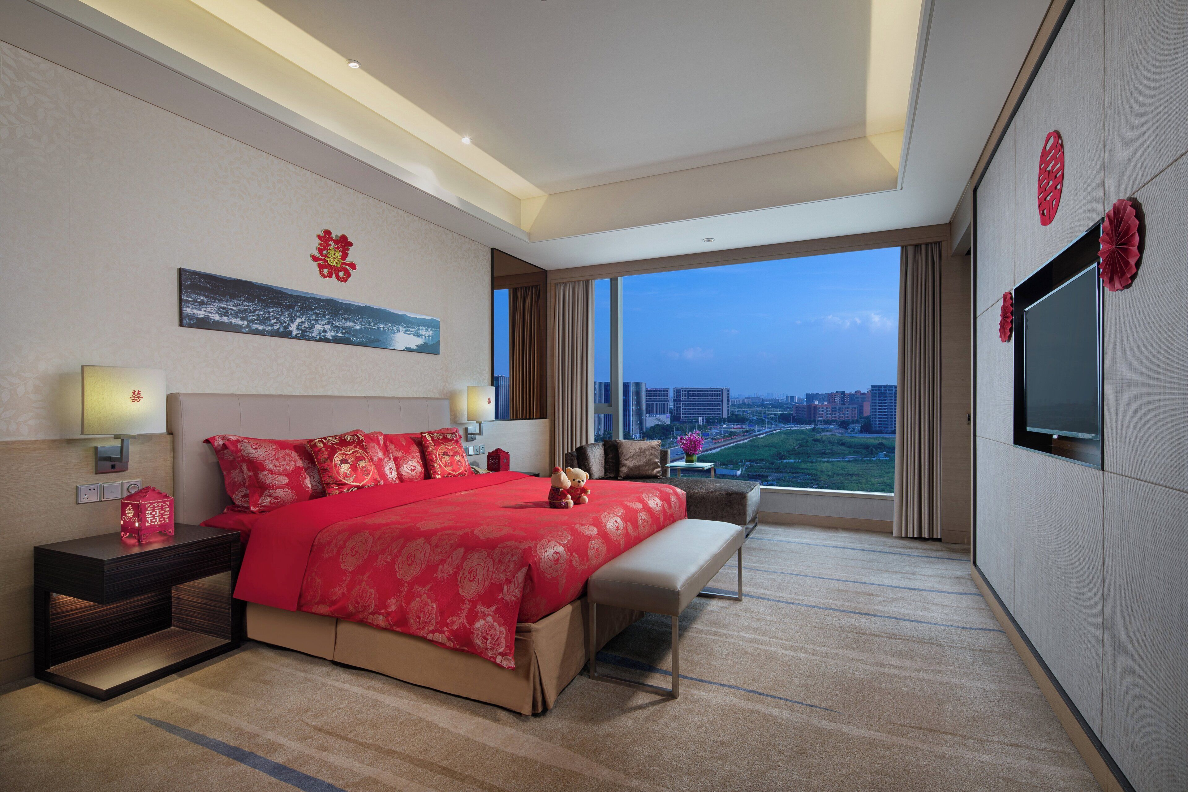 A hotel room with a large bed, bedside tables, a TV, and a view of the city.