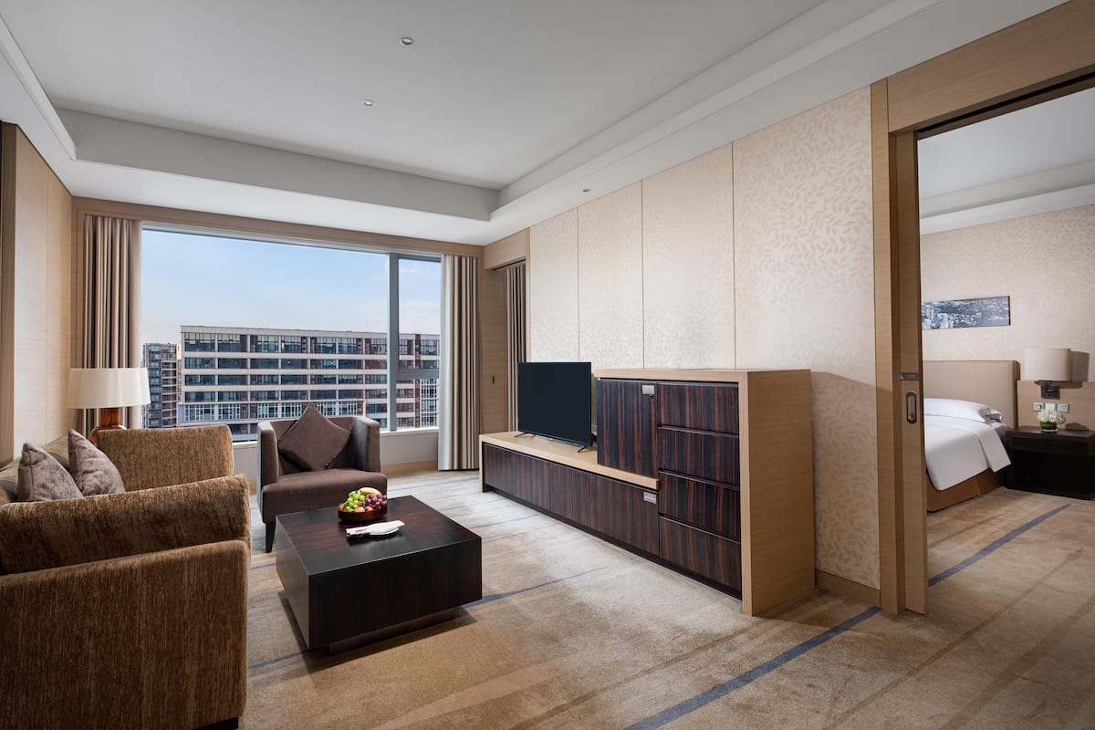A hotel room with a sofa, a coffee table, a TV, a wardrobe, a bed, and a view of the cityscape.