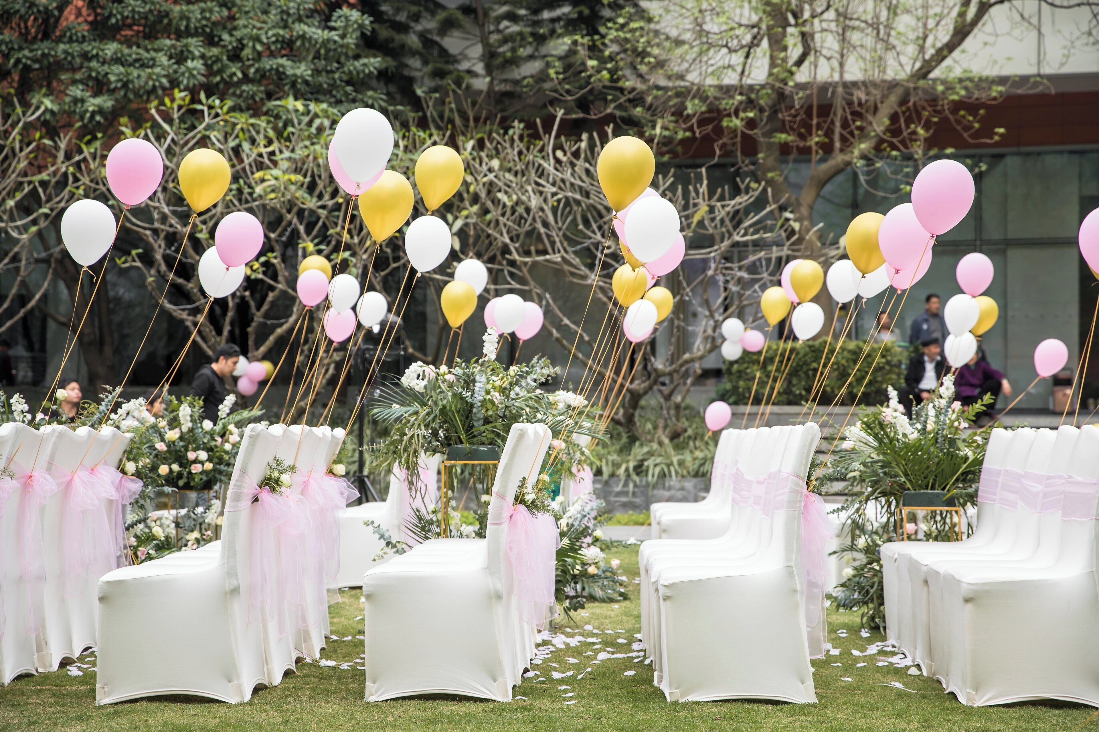 Sedute all'aperto con sedie bianche e nastri rosa, immerse nel verde e palloncini.