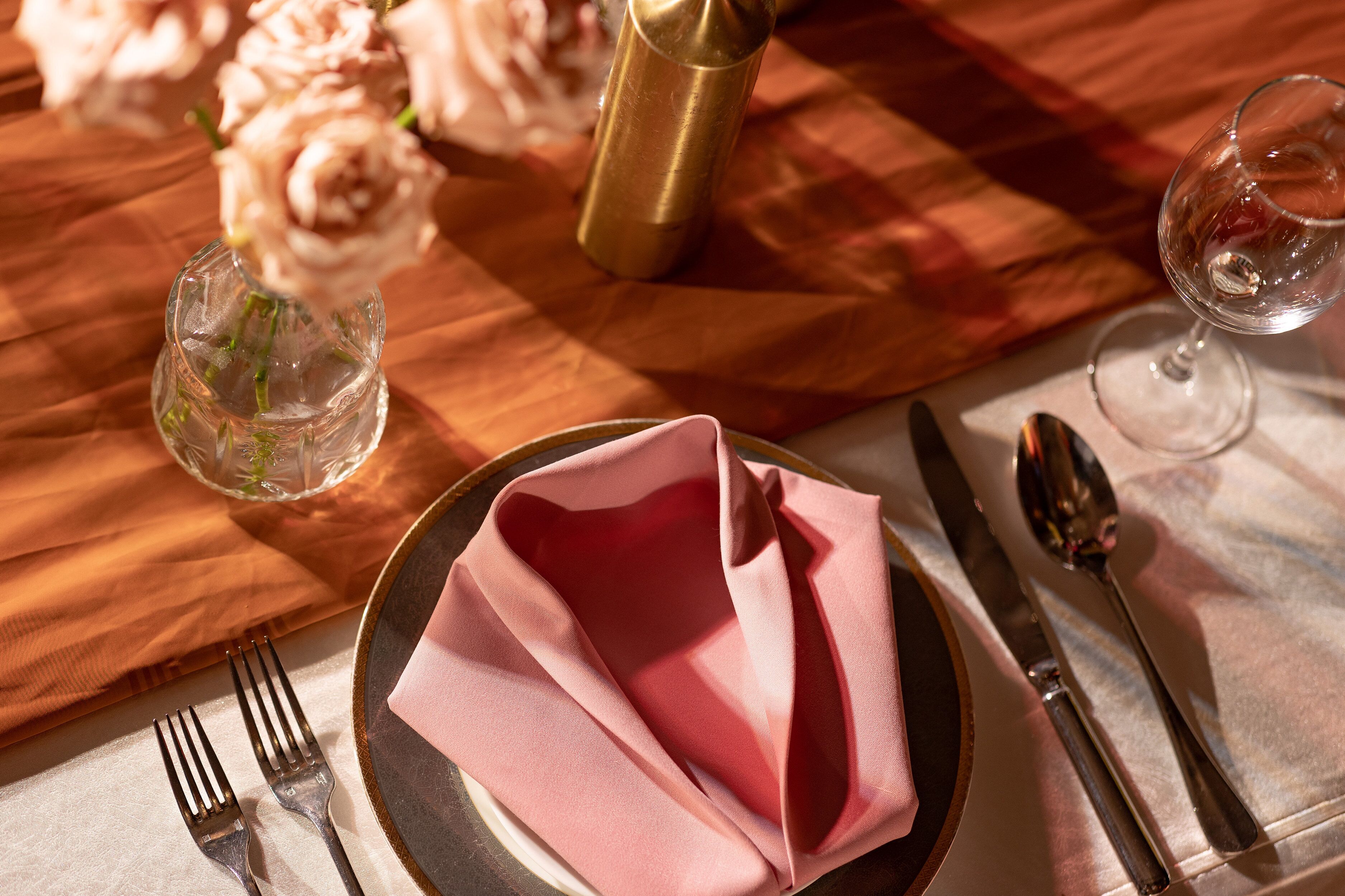 Mesa posta com guardanapo dobrado, um copo d'água, taça de vinho, talheres e um vaso com flores.