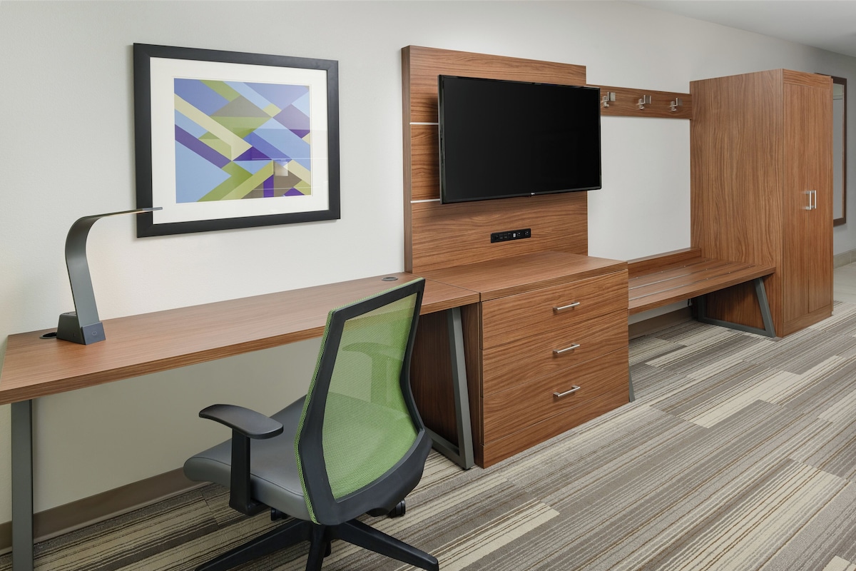 A modern office space with a long wooden desk, a green mesh office chair, a flat-screen TV mounted on the wall, and a large wooden wardrobe.