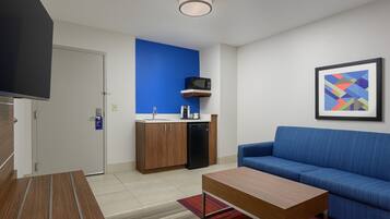 Una sala de estar moderna con un sofá azul, una mesa de centro de madera, una zona de cocina con microondas y fregadero, y un cuadro abstracto enmarcado en la pared.