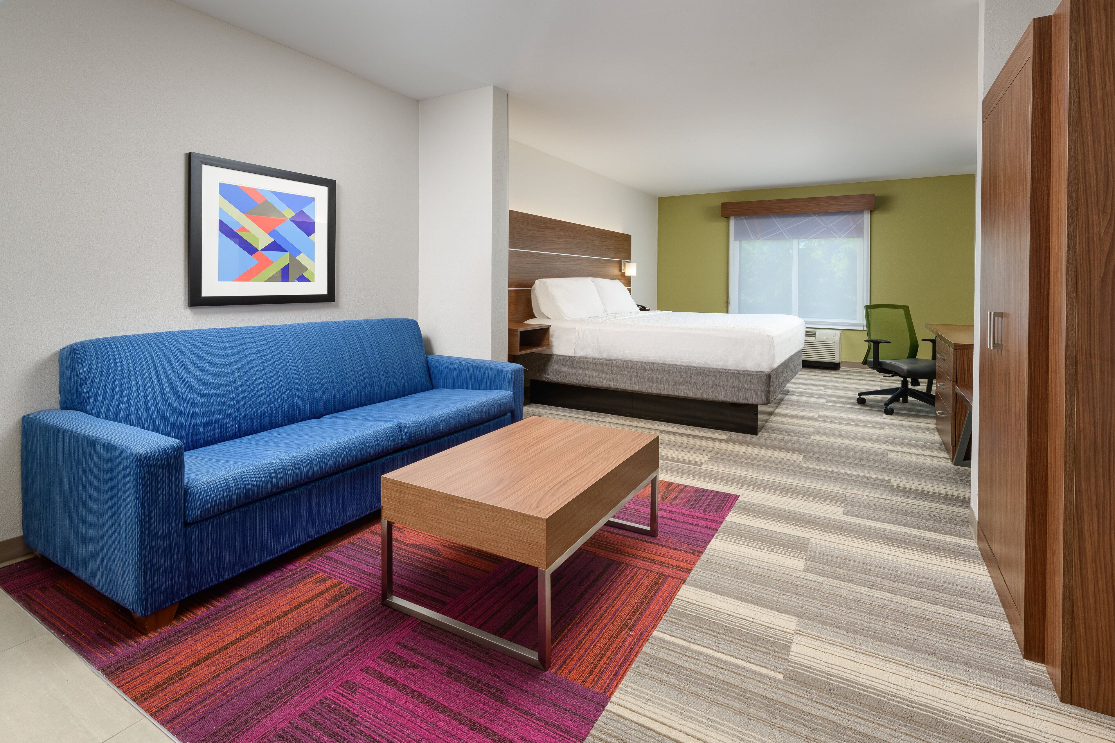 A hotel room with a blue sofa, a wooden coffee table, a bed with a white headboard, a desk with a chair, and a framed picture on the wall.
