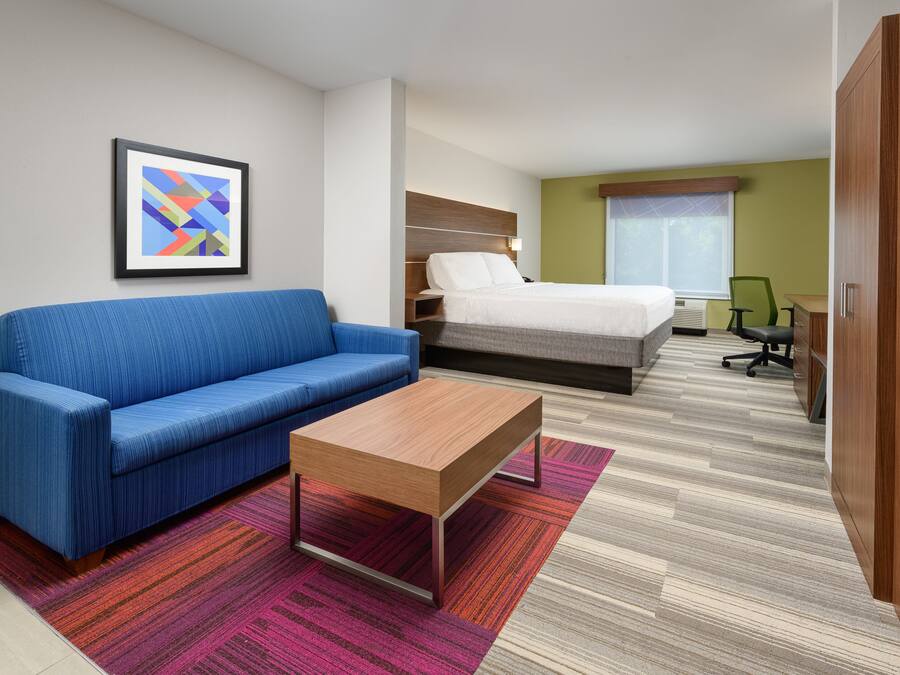 Une chambre d’hôtel avec un canapé bleu, une table basse en bois, un lit avec une tête de lit blanche, un bureau avec une chaise et un tableau encadré au mur.