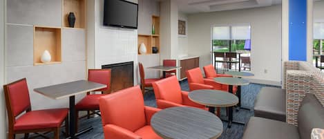 A hotel lobby with red chairs, round tables, a flat-screen TV, and a fireplace.