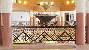 Un salón imponente con una gran lámpara de araña, columnas ricamente decoradas y una barandilla con diseño.