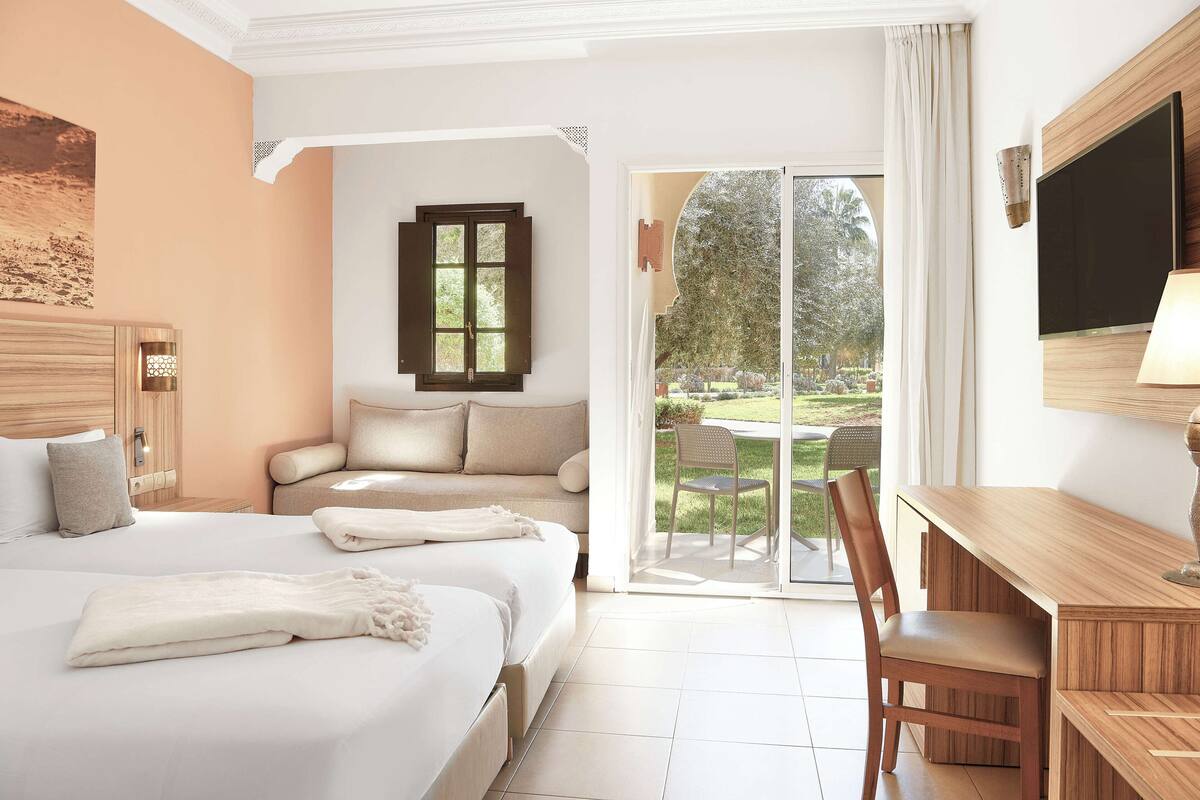 A hotel room with a bed, a sofa, a desk, and a TV. There is a sliding glass door leading to an outdoor area with a garden.