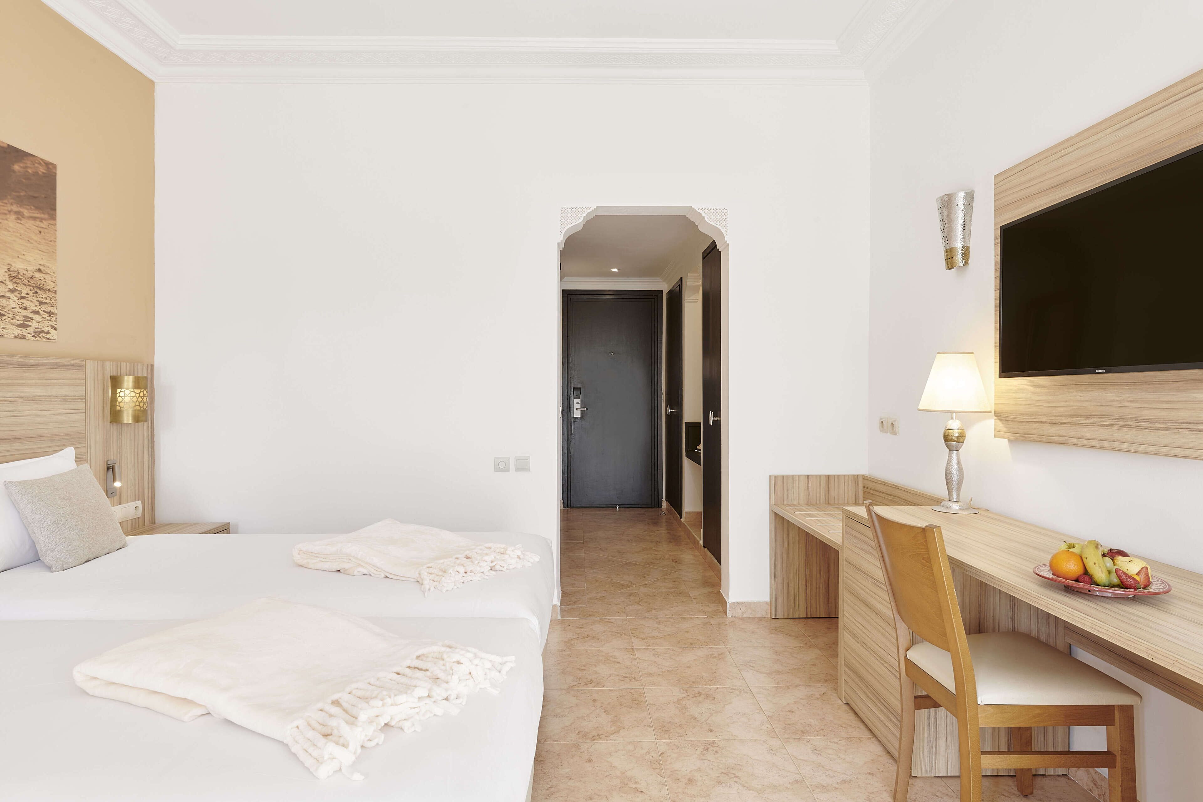 A hotel room with two beds, a desk with a chair, a television, and a fruit bowl.