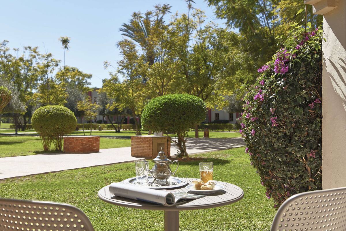 A table set outdoors with a pitcher, glasses, and a plate of food, surrounded by greenery and a clear sky.
