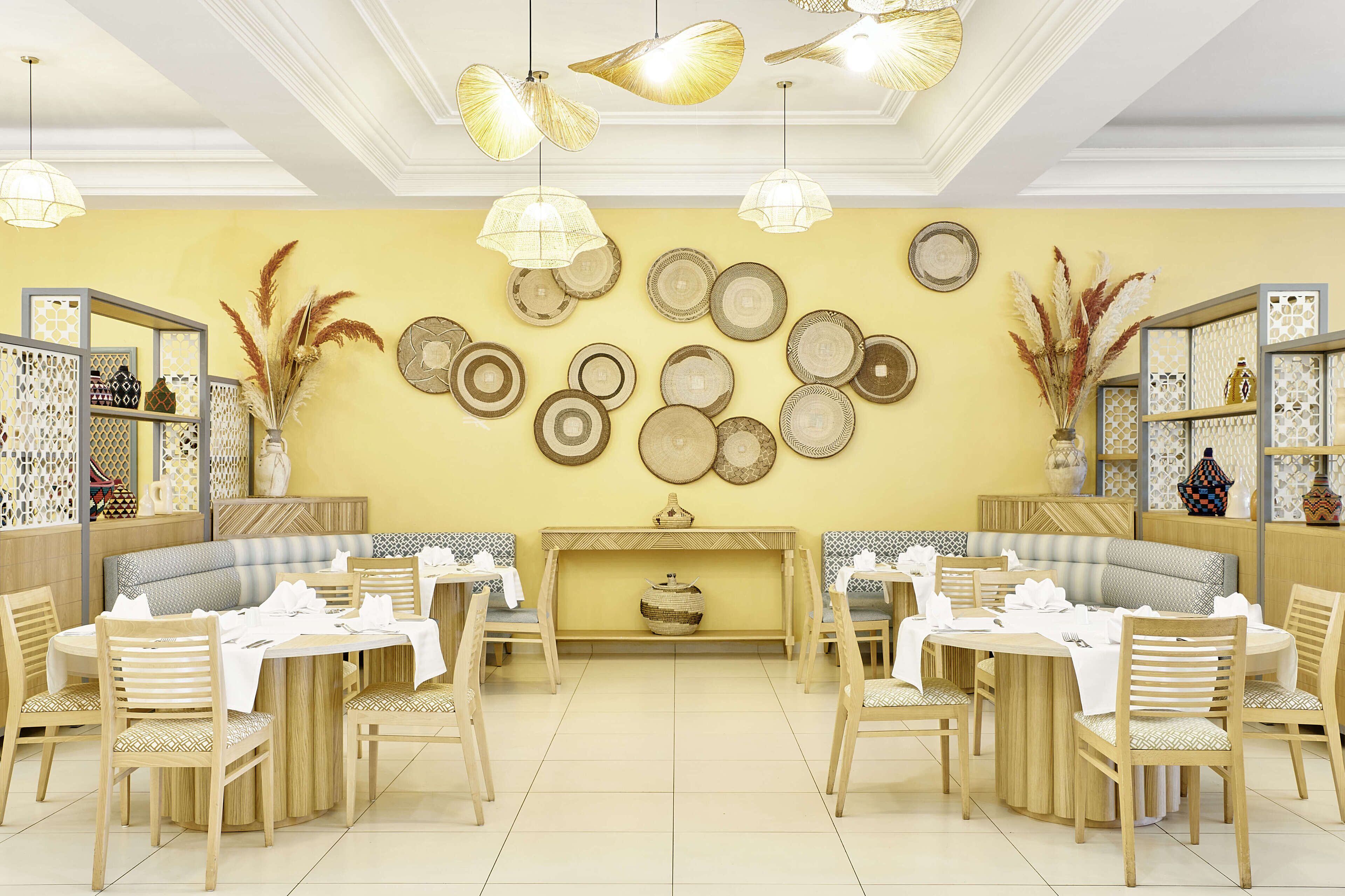 A restaurant with round tables, wooden chairs, and a yellow wall decorated with plates and vases.