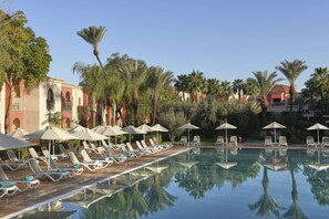 A poolside area with sun loungers and umbrellas, surrounded by palm trees and a building with arches. - Iberostar Waves Club Palmeraie Marrakech -All Inclusive (Marrakech)