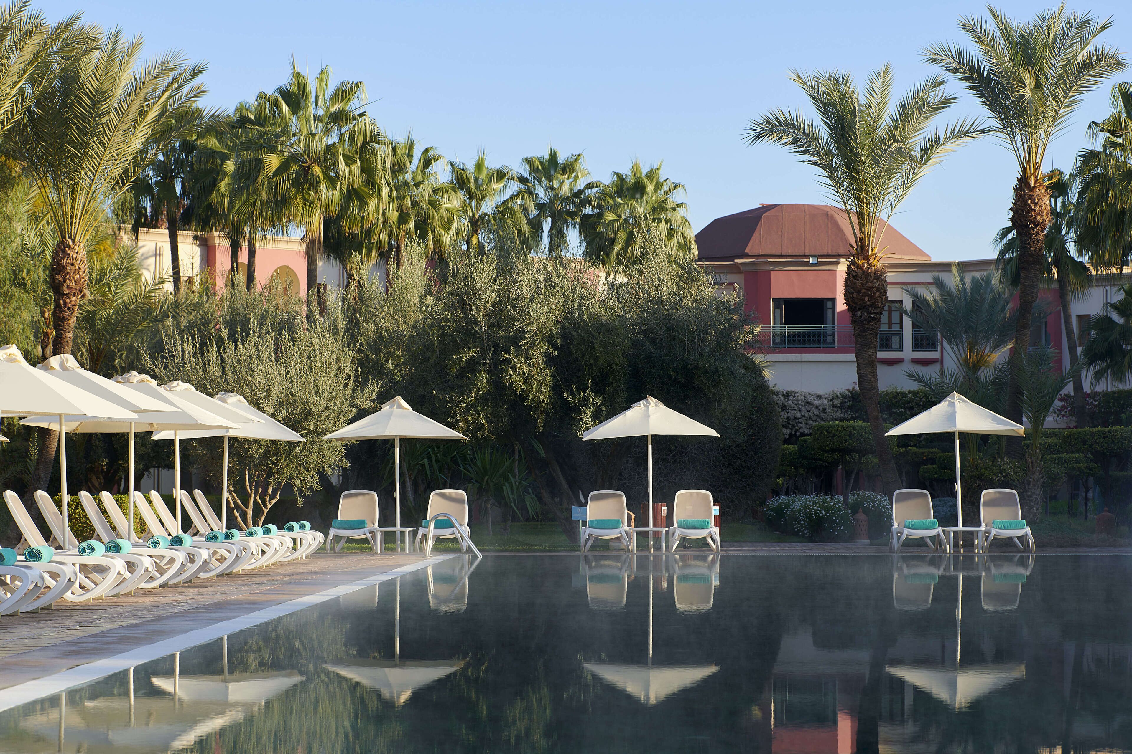 A pool area with lounge chairs and umbrellas, surrounded by palm trees and a building with a red roof.