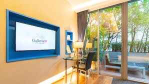 A hotel room with a large TV screen displaying the hotel name, a glass table with chairs, a sofa, and a view of greenery outside. - Galleria 10 Hotel Bangkok (Bangkok)