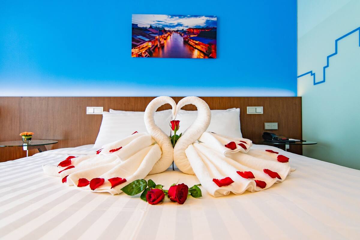 A neatly made bed with towels arranged in the shape of swans and a rose, with a cityscape painting above.