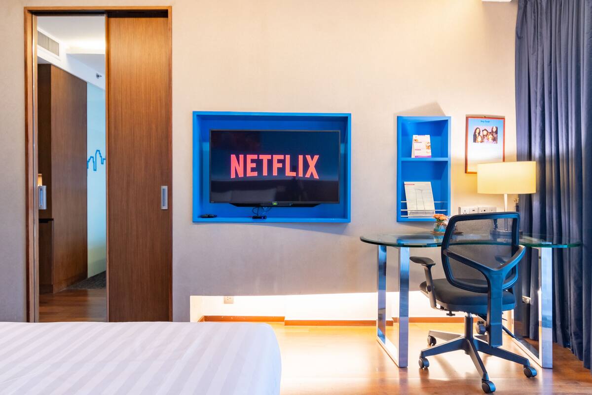 A hotel room with a bed, a desk with a chair, a TV mounted on the wall, and a framed picture on the wall.