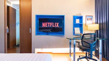 A hotel room with a bed, a desk with a chair, a TV mounted on the wall, and a framed picture on the wall.