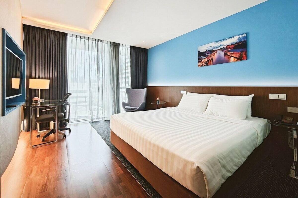 A hotel room with a large bed, a desk with a chair, a desk lamp, a chair, a window with curtains, and a painting on the wall.