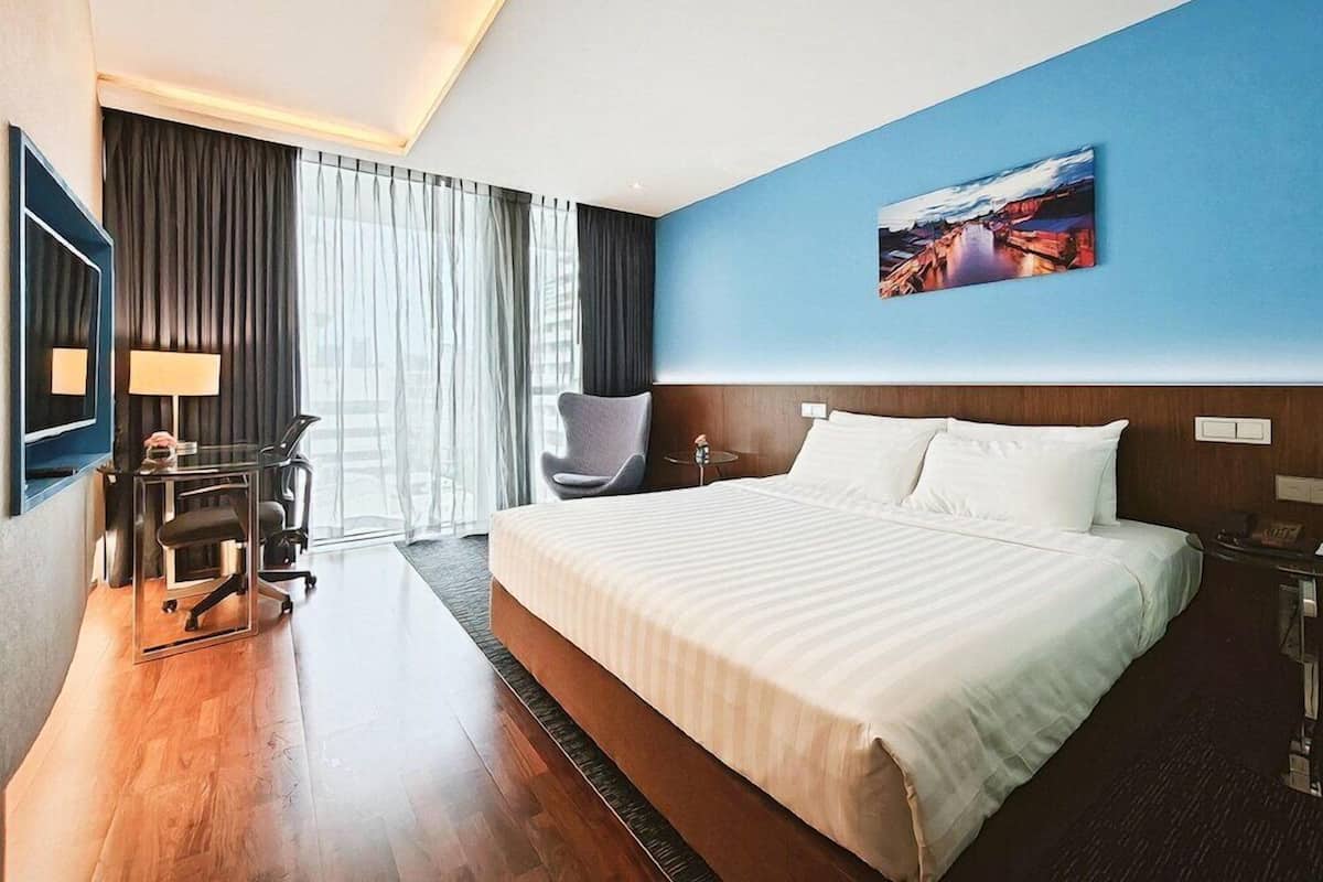 A hotel room with a large bed, a desk with a chair, a desk lamp, a chair, a window with curtains, and a painting on the wall.