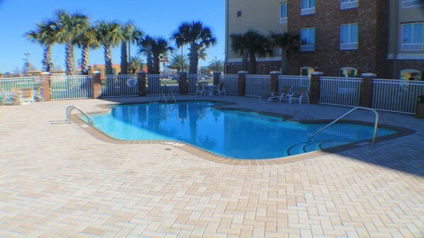 A rectangular swimming pool with a tiled deck, surrounded by a white metal fence and palm trees.
