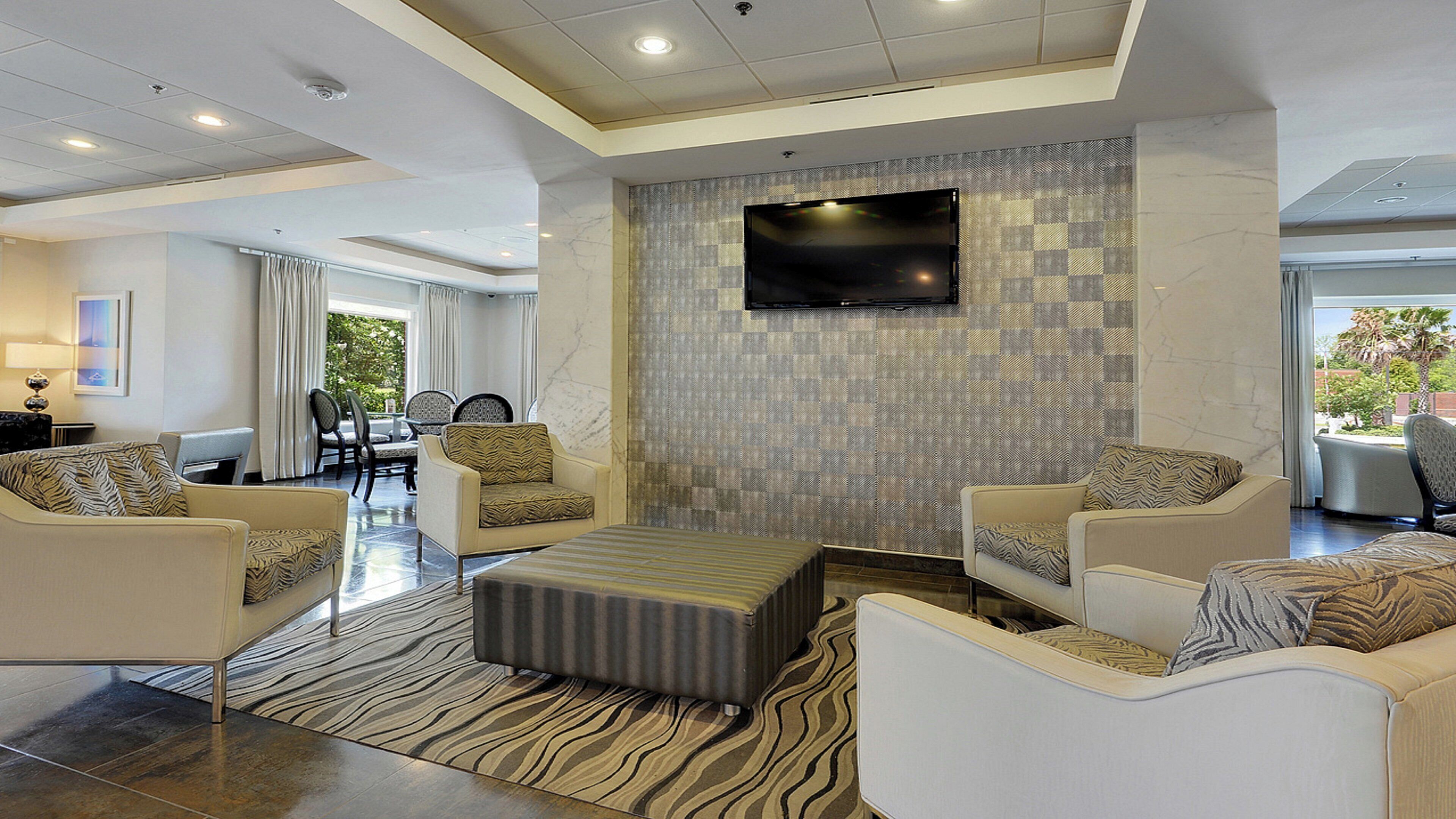 A modern hotel lobby with a flat-screen TV mounted on the wall, a seating area with sofas and chairs, and a patterned rug on the floor.