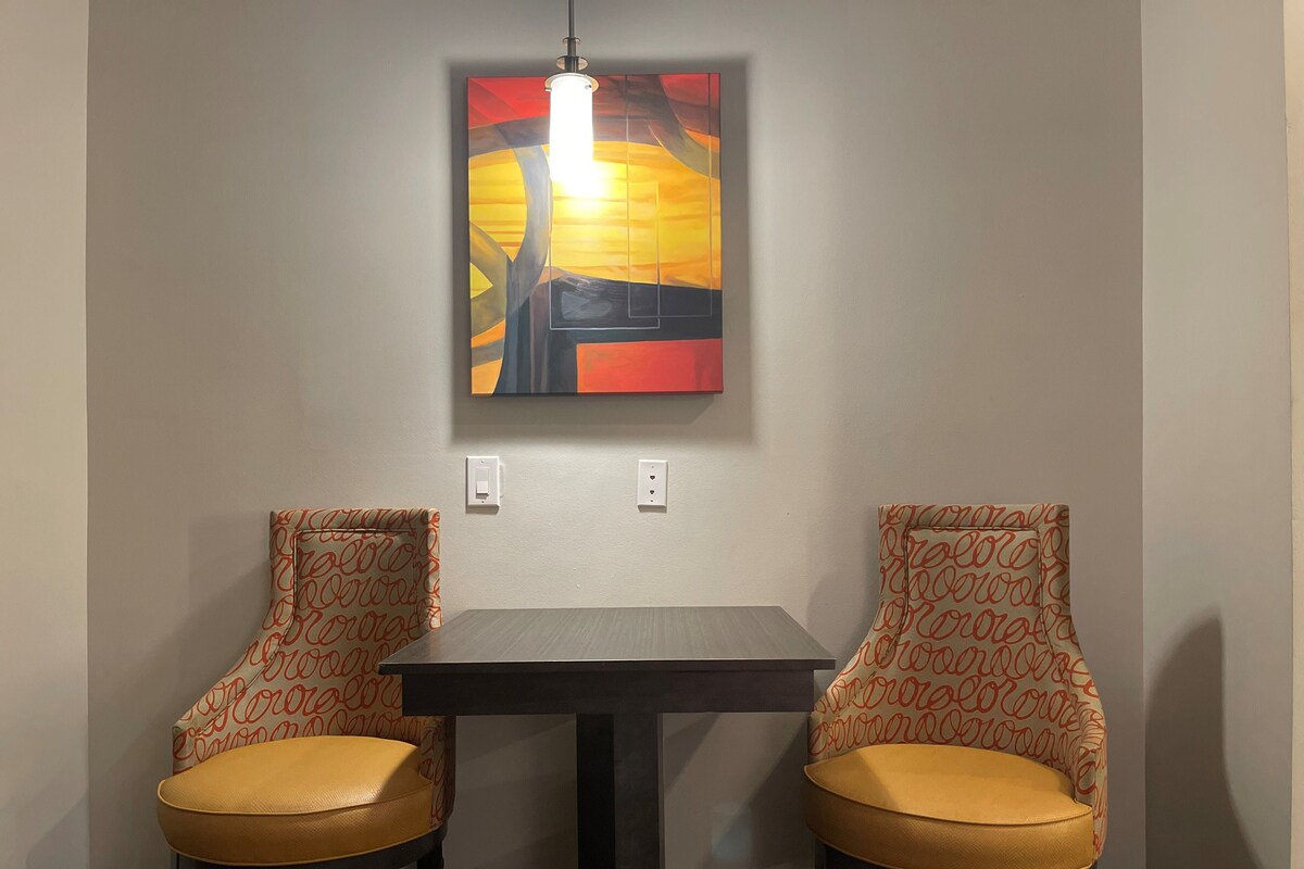 Two chairs with patterned upholstery and yellow cushions, a table, and a wall-mounted painting above them.