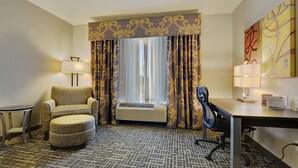 Ein Hotelzimmer mit Sofa, Ohrensessel, Hocker, Schreibtisch und Klimaanlage.