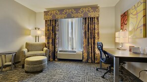 A hotel room with a sofa, armchair, ottoman, desk, and air conditioning unit.