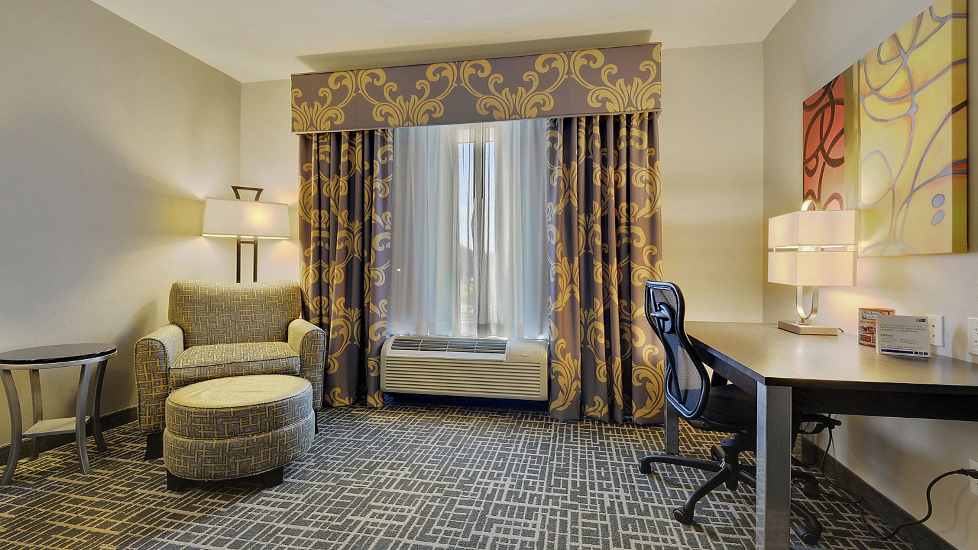 Habitación de hotel con sofá, sillón, reposapiés, escritorio y un ventanal con cortinas.