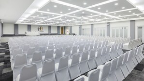 Un salón de conferencias con filas de sillas blancas, un escenario con un atril y un logotipo grande en la pared.