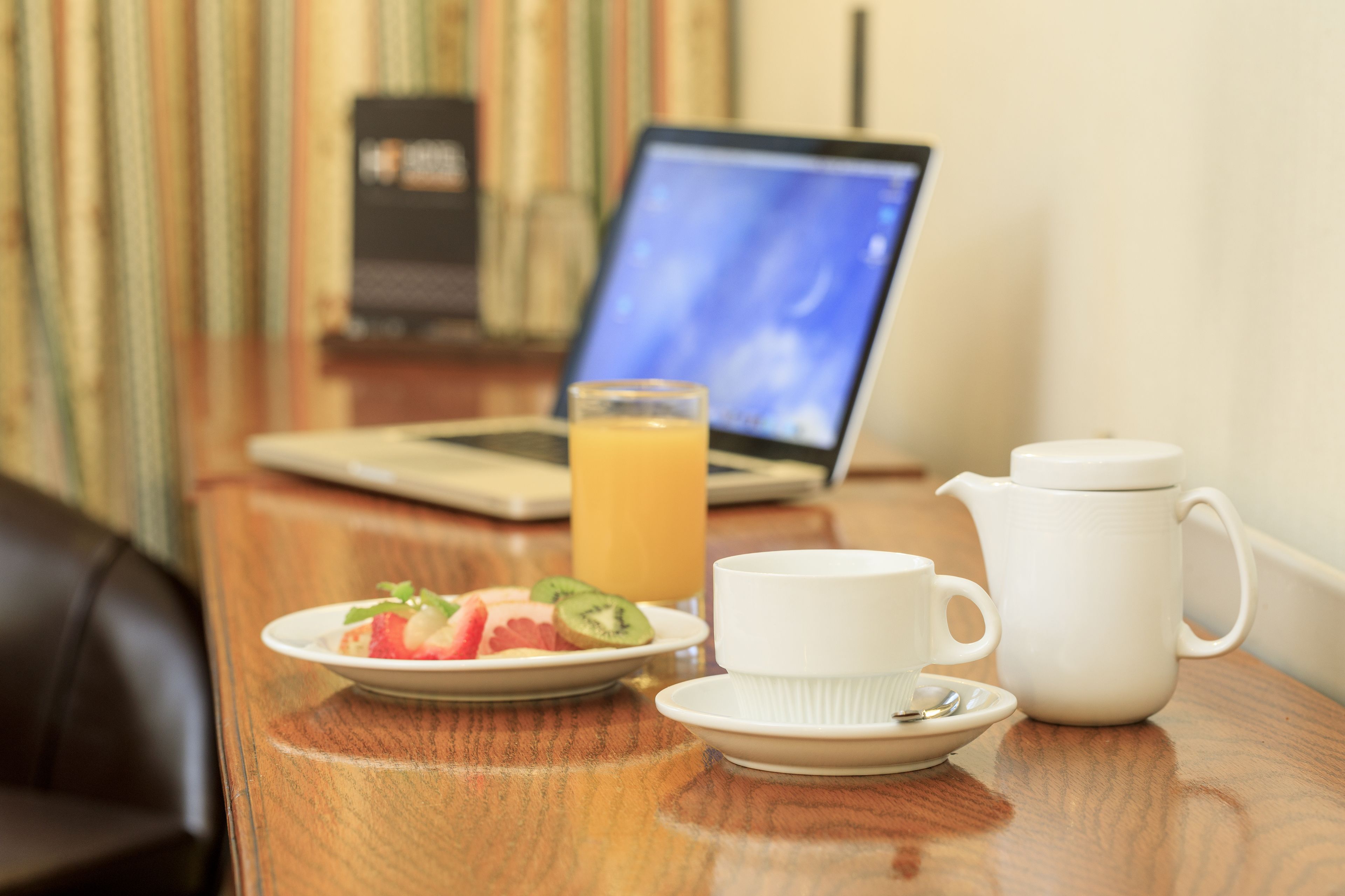 A breakfast setup with a laptop, a glass of orange juice, a white teapot, and a plate with fruit on a wooden table.