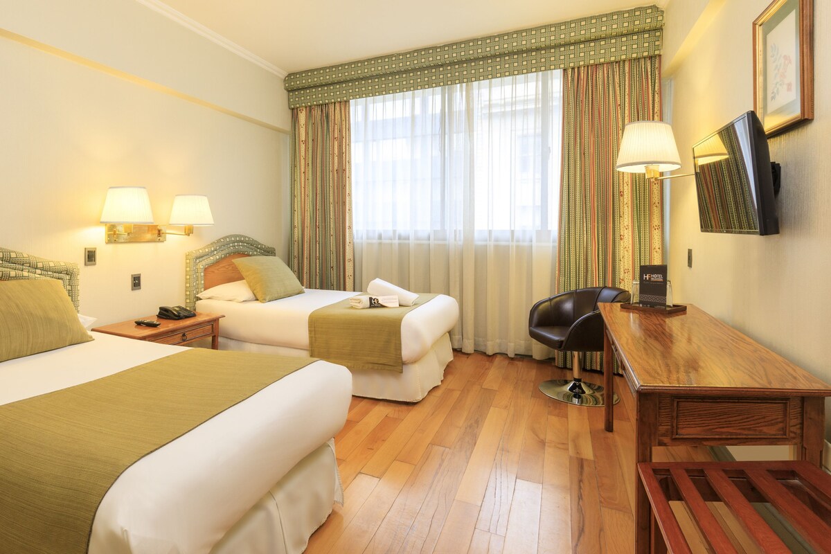 A hotel room with two beds, a desk, a chair, a TV, and a window with curtains.