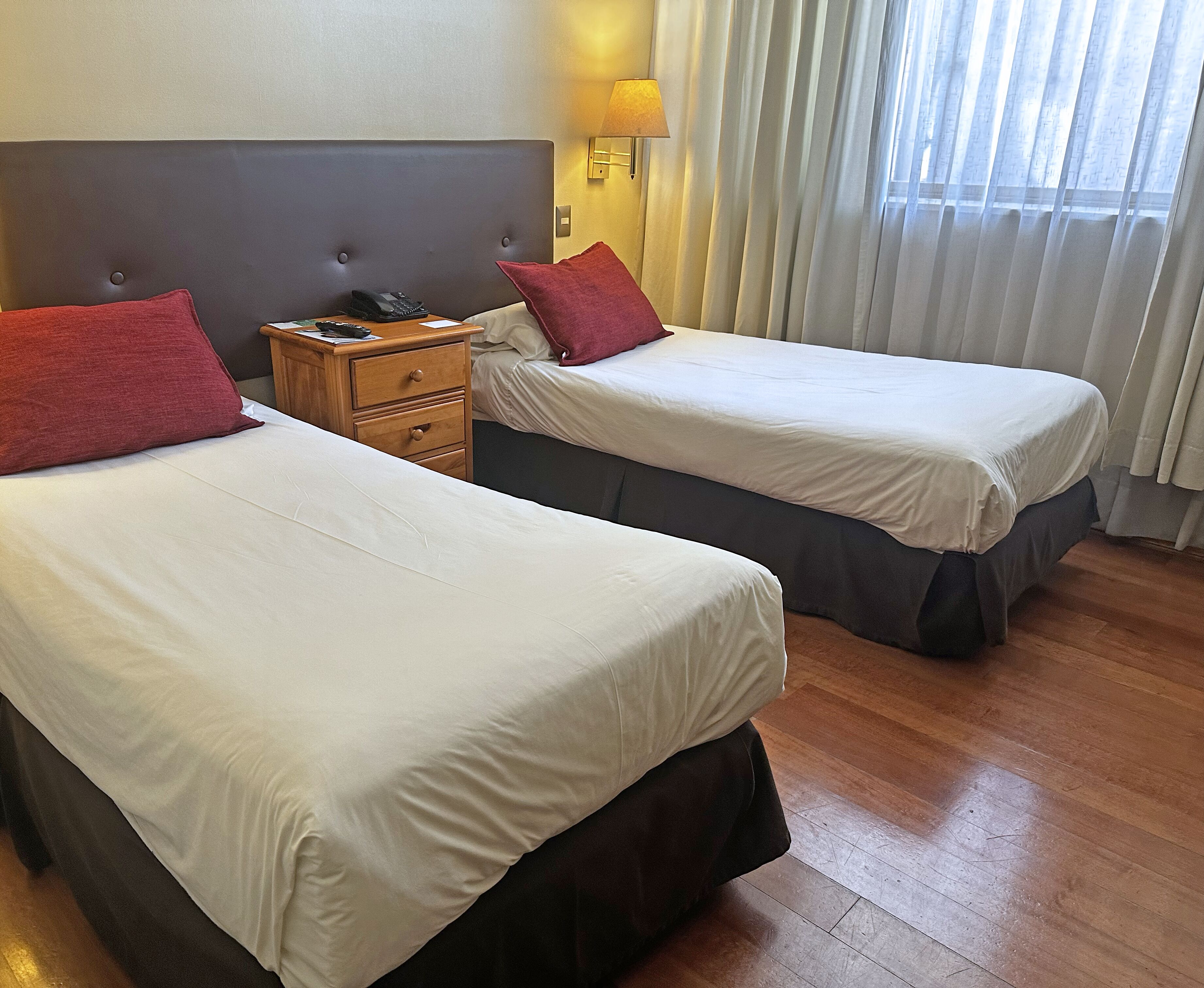 Two single beds with white linens and red pillows, a wooden nightstand with a phone, and a lamp.