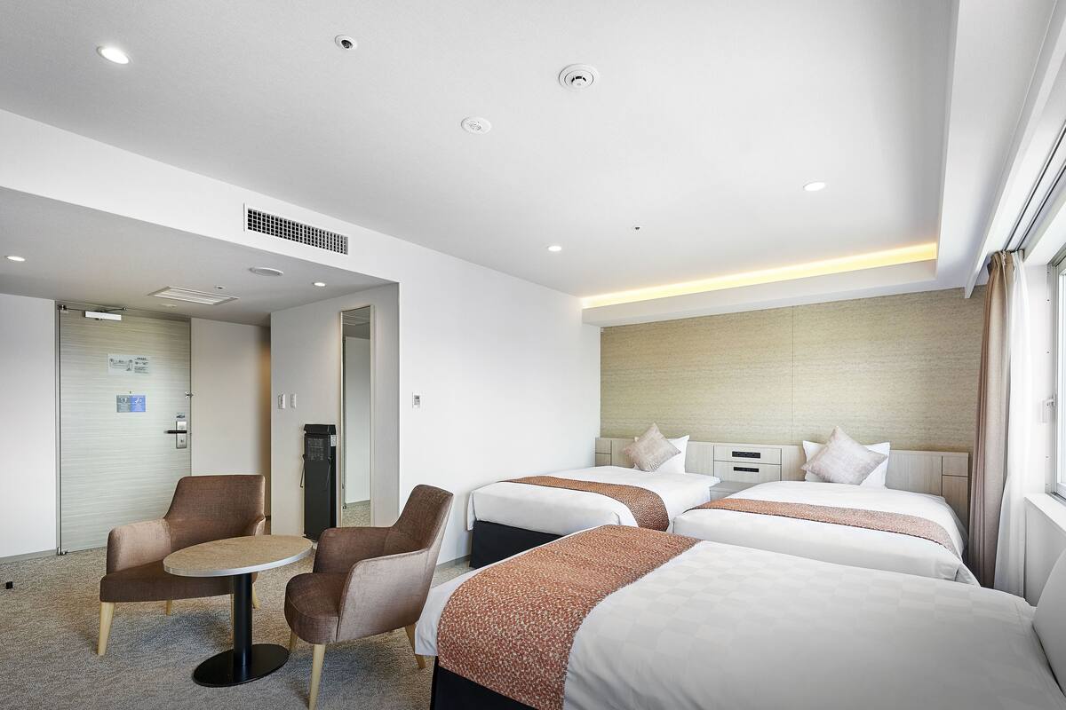 Ein Hotelzimmer mit drei Betten, einem kleinen Tisch, zwei Sesseln und einem Fenster mit Vorhängen.