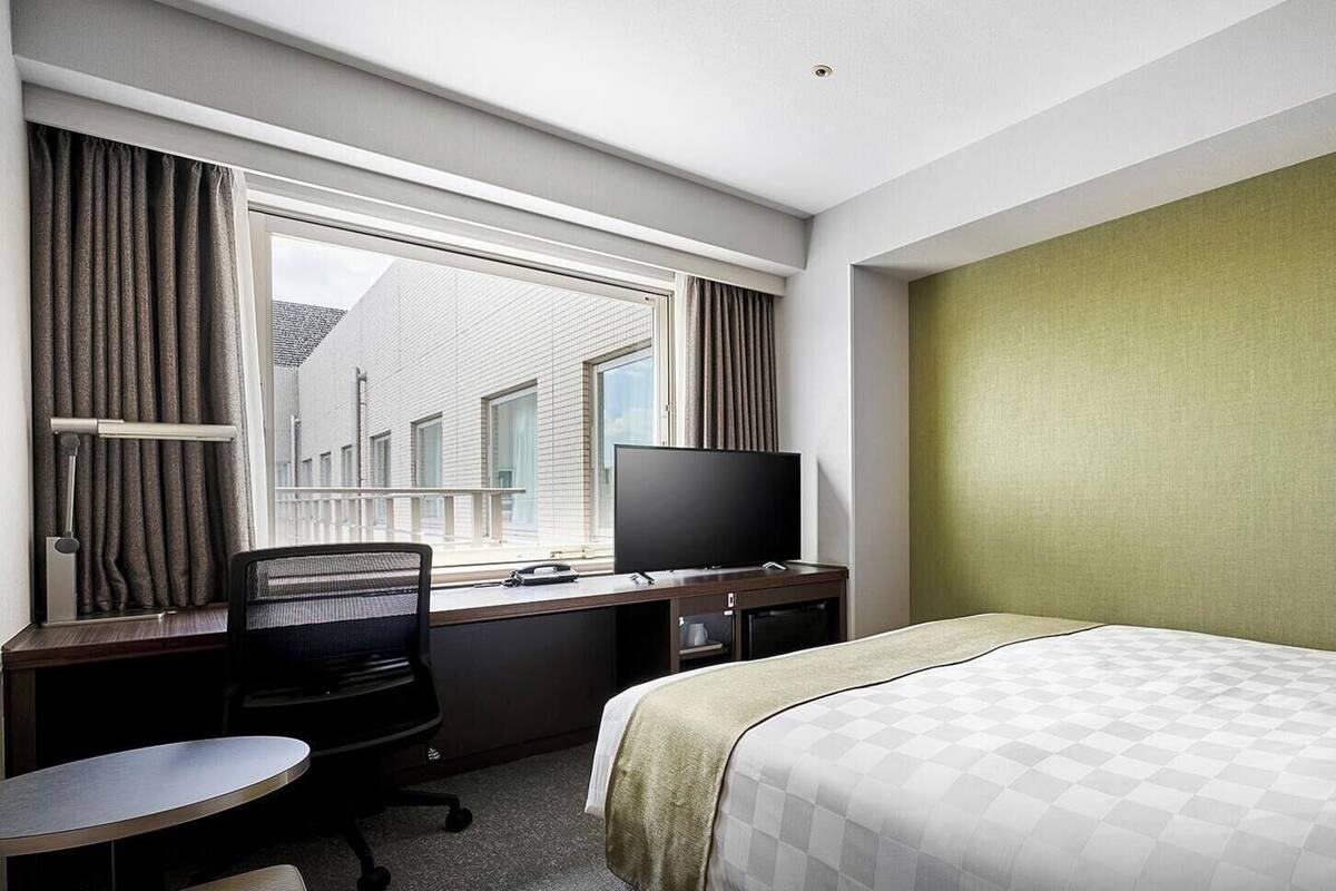 Ein Hotelzimmer mit Bett, Schreibtisch mit Computer, Fernseher und einem Fenster mit Vorhängen.