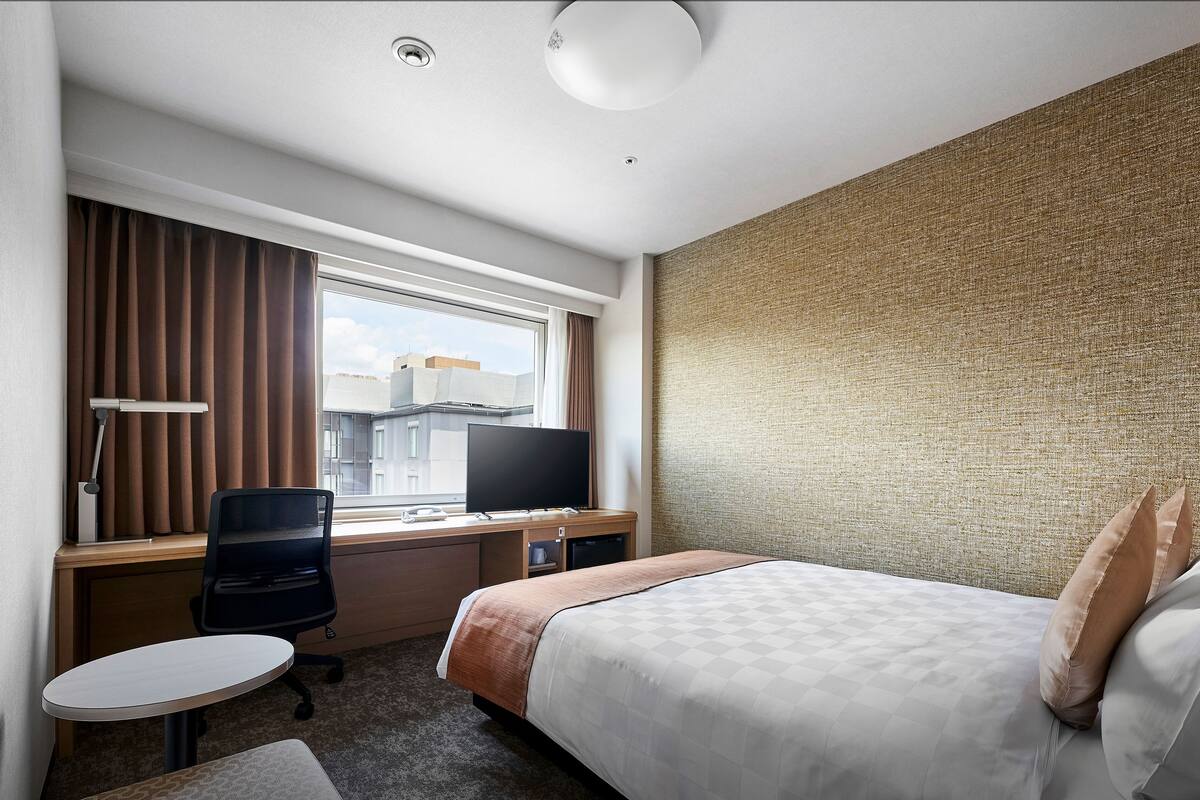 Ein Hotelzimmer mit einem Bett, einem Schreibtisch mit Computer, einem Stuhl, einem kleinen Tisch und einem Fenster mit Vorhängen.