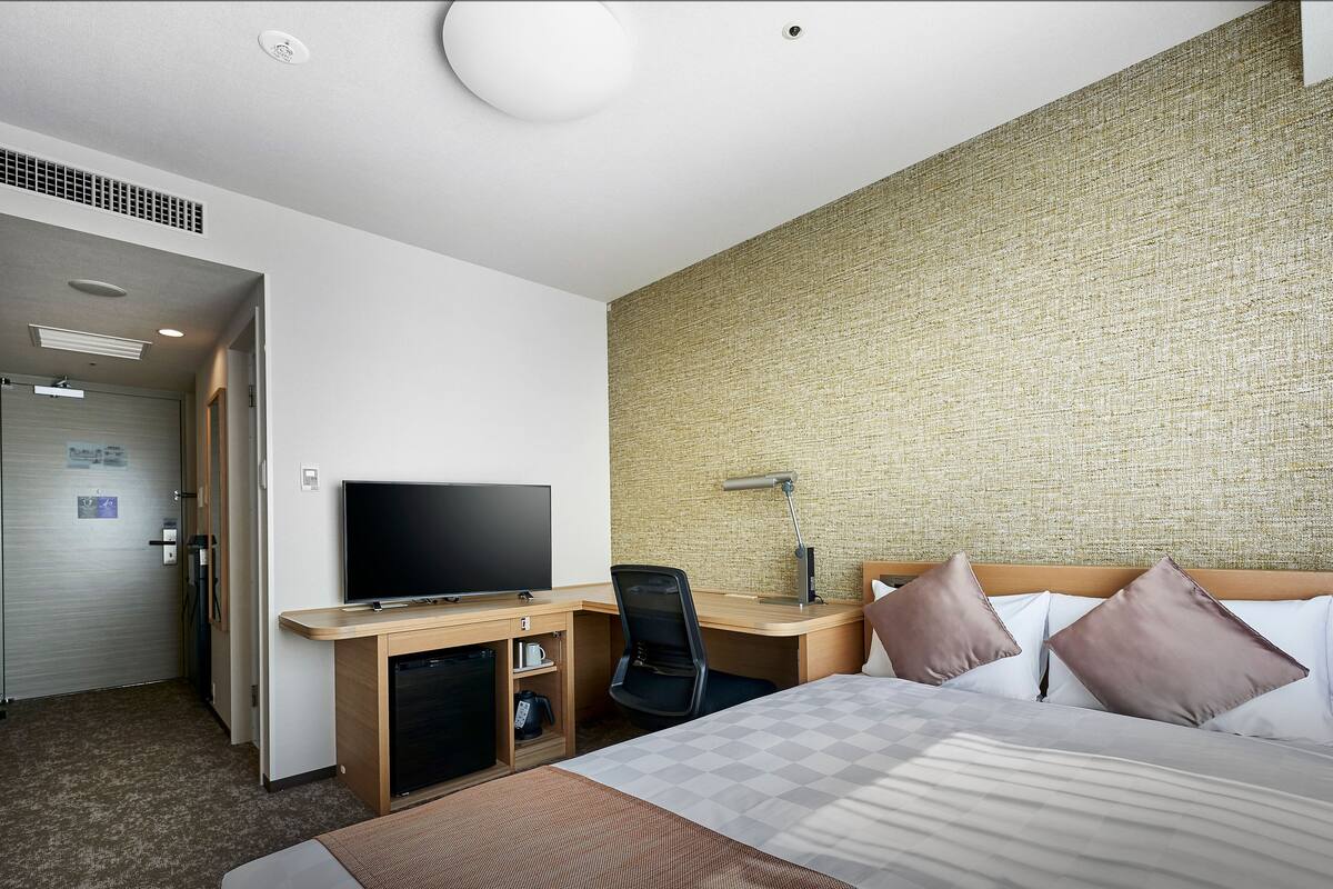 Ein Hotelzimmer mit einem Bett, einem Schreibtisch mit Fernseher, einem Sessel, einer Schreibtischlampe und einer Wand mit strukturierter Oberfläche.