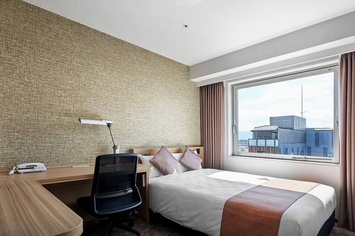 Ein Hotelzimmer mit großem Fenster, einem Bett, einem Schreibtisch mit Stuhl, einem Fernseher und einem an der Wand montierten Telefon.
