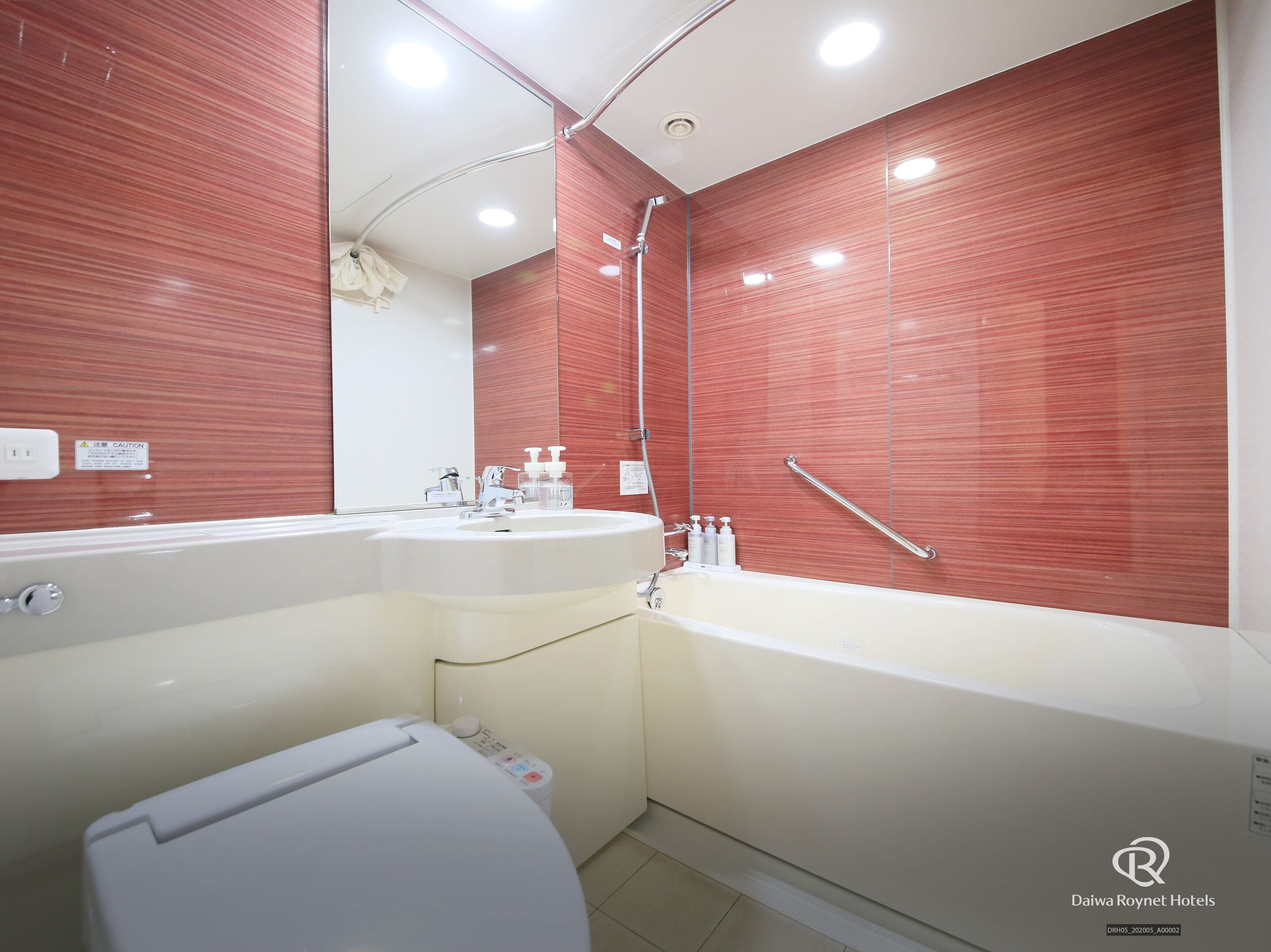A modern bathroom with a white sink, a bathtub, and a toilet. There are hand sanitizer bottles on the sink and a handrail on the wall.