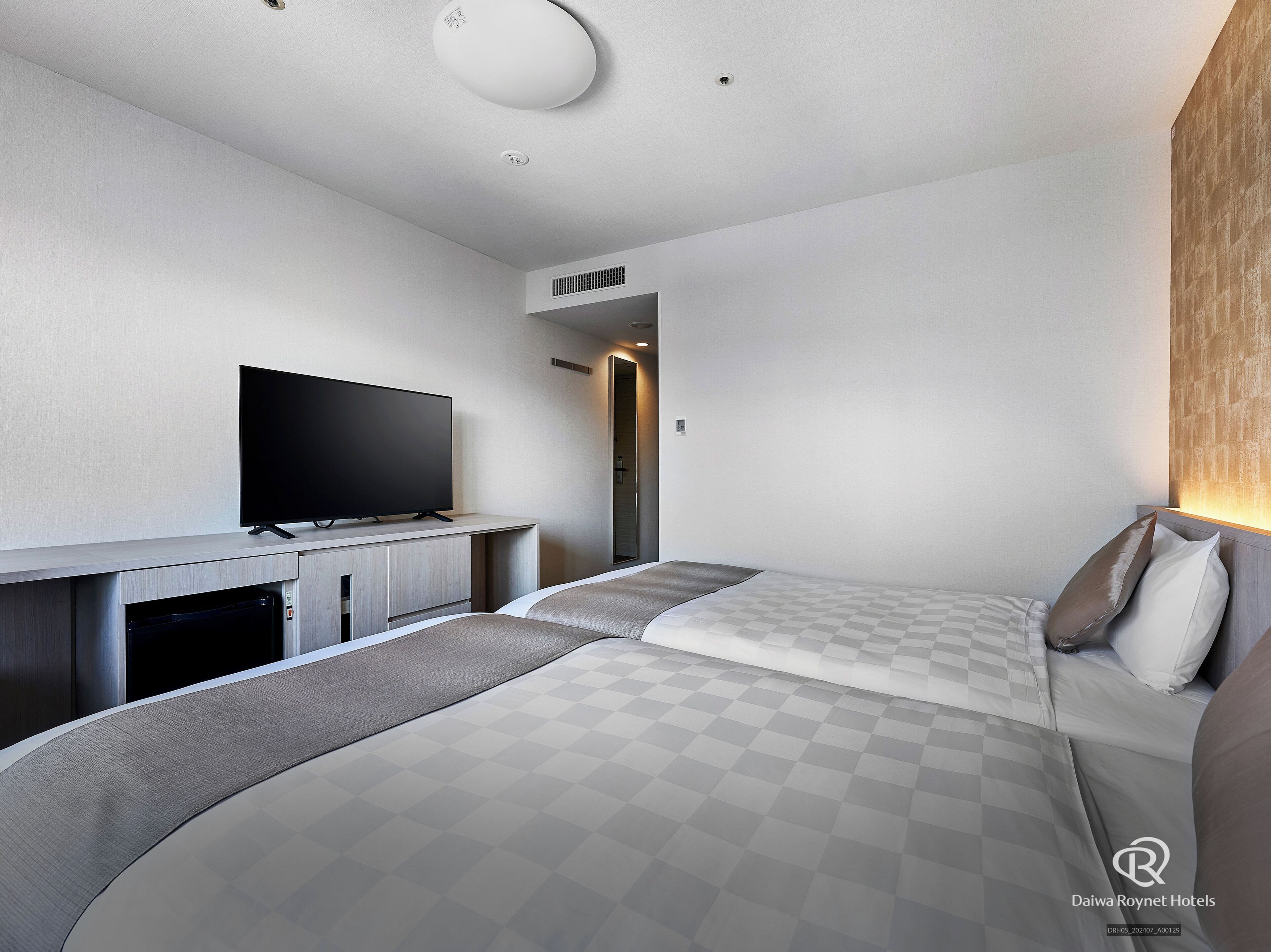 Una habitación de hotel moderna con televisión de pantalla plana, una cama con ropa de cama a cuadros, un escritorio pequeño y un calefactor integrado.