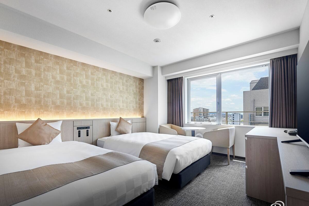 Ein Hotelzimmer mit zwei Betten, einem Schreibtisch, einem Stuhl und einem großen Fenster mit Blick auf die Stadt.