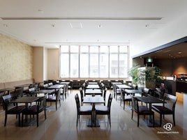 A spacious dining area with multiple tables and chairs, large windows, and a kitchen area.