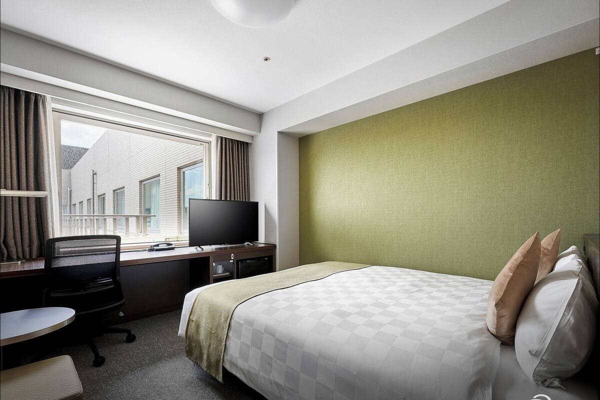 Ein Hotelzimmer mit einem Bett, einem Schreibtisch mit Stuhl, einem Fernseher und einem Fenster mit Vorhängen.