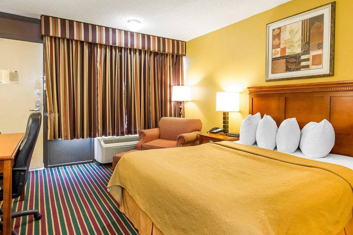 A hotel room with a bed, a desk, an air conditioner, a chair, a lamp, and a painting on the wall.
