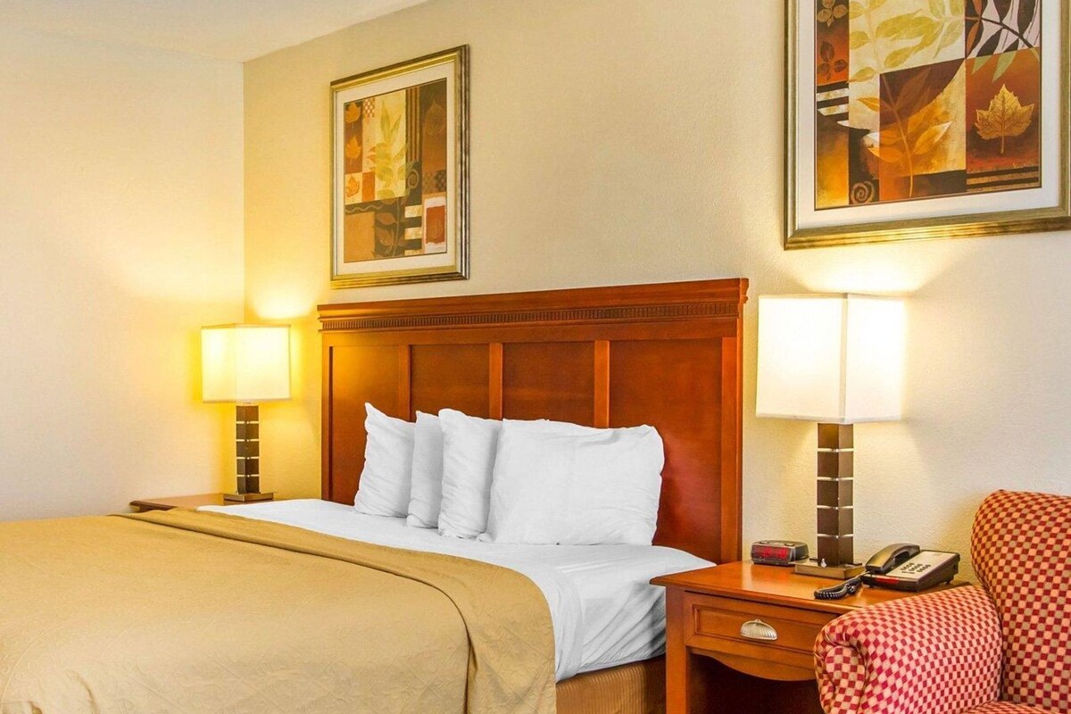 Een hotelkamer met een bed, bedlampen, een stoel, een nachtkastje en twee kunstwerken in een lijst aan de muur.