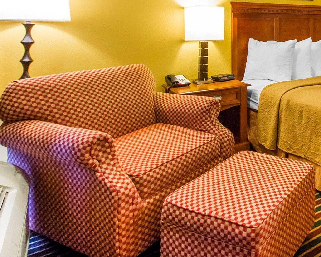 Habitació d'hotel amb una butaca estampada de quadradets, un otomà a joc, una taula de nit amb una làmpada, un telèfon i un llit amb nujols blancs.
