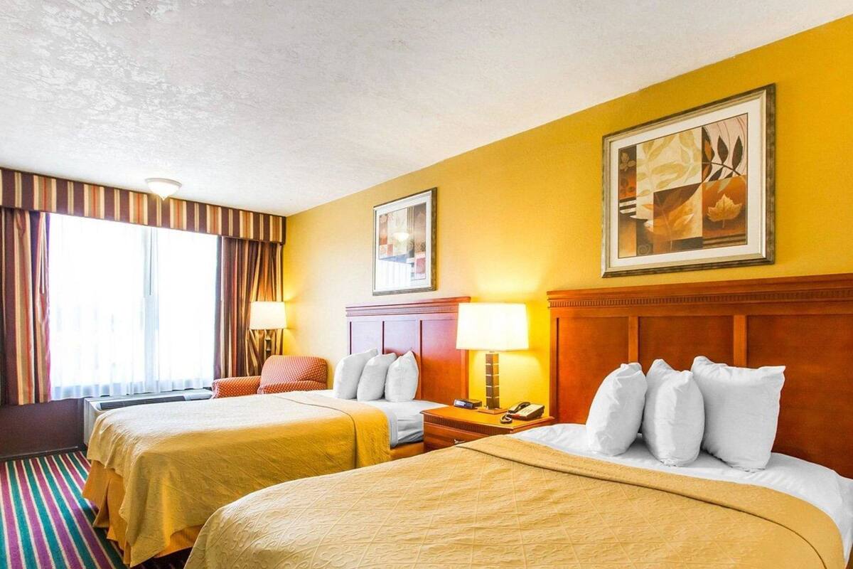 A hotel room with two beds, a large window with curtains, a painting on the wall, and a striped carpet.