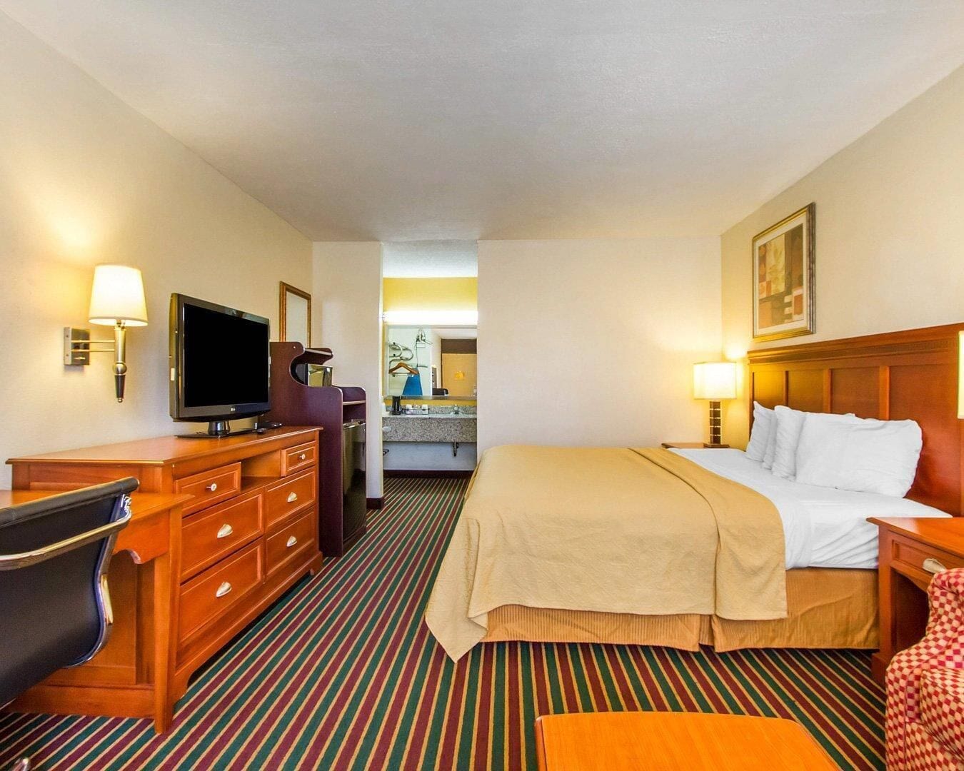 A hotel room with a bed, a desk with a television, a chair, a dresser, and a bathroom visible through a door.