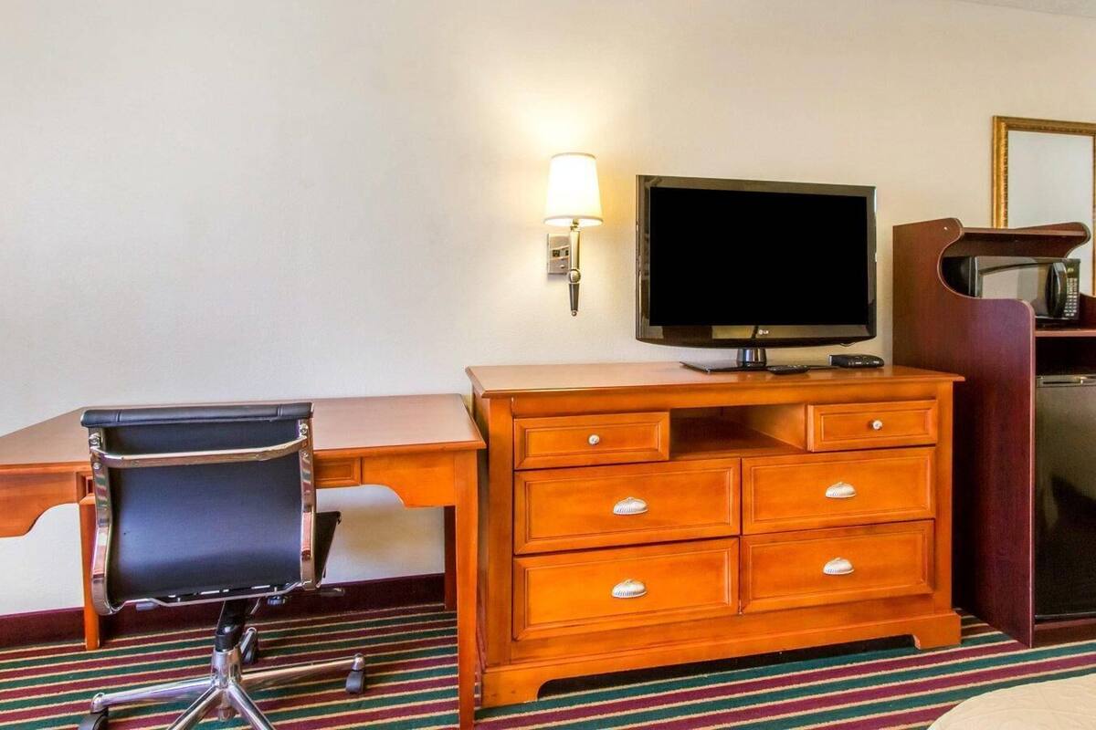 A hotel room with a wooden desk, a black office chair, a television, a lamp, and a microwave.