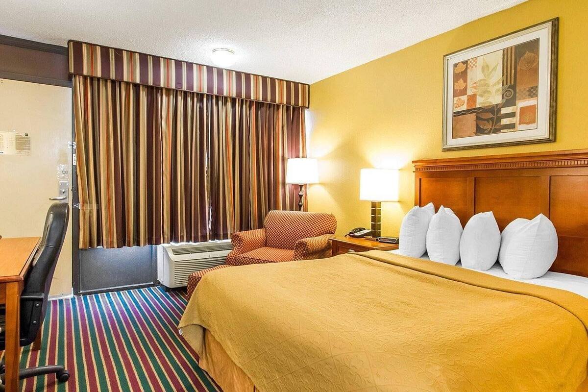 A hotel room with a bed, a desk, a chair, a lamp, and a painting on the wall.